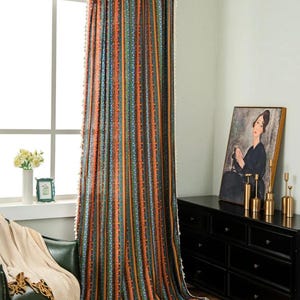 Può includere: Una tenda colorata con strisce verticali nei toni dell'arancione, blu, verde e marrone. La tenda ha un motivo strutturato ed è rifinita con frange bianche sui bordi. La tenda è appesa davanti a una finestra.