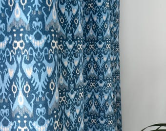 May include: Blue patterned curtain with a repeating ikat design in shades of blue and white. The fabric is folded, showing the texture and pattern. The curtain is hanging in front of a white wall.