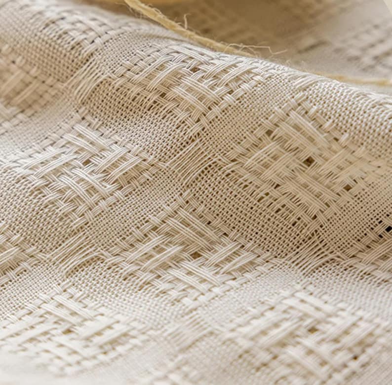 May include: Close-up of a woven fabric with a textured, basket-weave pattern. The fabric is a neutral beige color, with visible threads creating a grid-like design. A piece of twine is visible in the upper portion of the image.