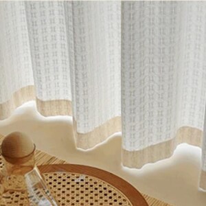 May include: Off-white textured curtains with a beige border. The curtains have a repeating square pattern and are partially drawn, revealing a glimpse of a decorative table setting with a glass bottle and a woven tray.