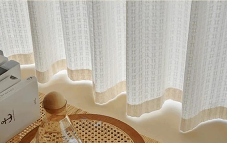May include: Off-white textured curtains with a beige border. The curtains have a repeating square pattern and are partially drawn, revealing a glimpse of a decorative table setting with a glass bottle and a woven tray.