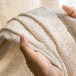 May include: Close-up of a folded, off-white woven fabric. The fabric has a textured, grid-like pattern. The material appears soft and lightweight, with visible details in the weave. The fabric is held in place by a person's hand.