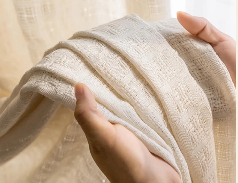 May include: Close-up of a folded, off-white woven fabric. The fabric has a textured, grid-like pattern. The material appears soft and lightweight, with visible details in the weave. The fabric is held in place by a person's hand.