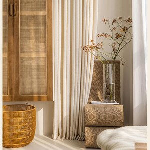 May include: A room interior featuring a wooden cabinet with woven panels, a woven basket, and cream-colored curtains. A tall glass vase holds a branch with berries, resting on stacked textured blocks.