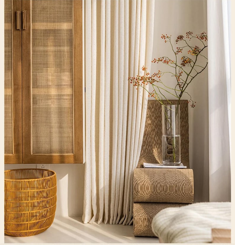 May include: A room interior featuring a wooden cabinet with woven panels, a woven basket, and cream-colored curtains. A tall glass vase holds a branch with berries, resting on stacked textured blocks.