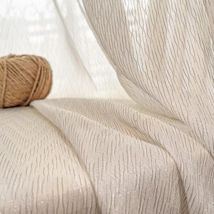 May include: Off-white sheer curtain fabric with vertical textured lines and subtle shimmer. A ball of natural twine sits in the background. The fabric drapes in soft folds, creating a delicate and airy aesthetic. Ideal for home decor and window treatments.