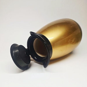 May include: A gold-colored, egg-shaped container with a black rim and lid. The lid is open, revealing the interior. The container has a glossy finish and is set against a white background. The container is likely a decorative item or a small urn.