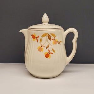 May include: Cream-colored ceramic pitcher with a lid, featuring a floral design in orange, yellow, and brown. The pitcher has a curved handle and a spout. The lid is topped with a small, pointed finial. The pitcher has vertical ribbed detailing.