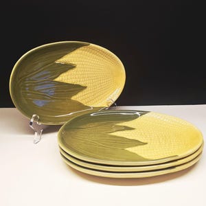 May include: Set of ceramic plates with a corn-on-the-cob design. The plates feature a yellow center with a textured pattern, and green leaves on the side. One plate is displayed on a clear stand, while the others are stacked.
