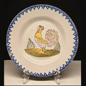 May include: A white ceramic plate with a hand-painted rooster design. The rooster is yellow, orange, and white, standing on a brown base with green foliage. The plate has a blue sawtooth border.