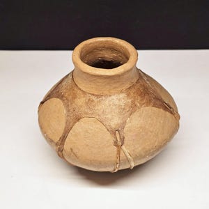 May include: A handmade, small, tan-colored ceramic pot with a rounded body and a narrow neck. The pot features a decorative pattern of lighter tan shapes outlined with darker brown lines, adding a rustic aesthetic. The pot is suitable for home decor.