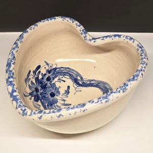 May include: Heart-shaped ceramic bowl with a cream-colored interior and a blue floral design. The rim is decorated with a blue speckled pattern. The bowl is a decorative piece for serving or display.