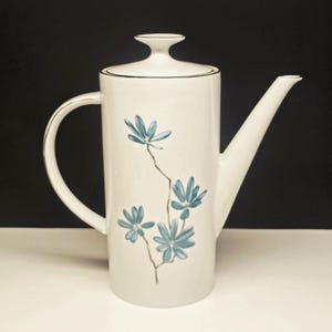 May include: A white ceramic coffee pot with a black rim and handle, featuring a floral design of blue flowers on a slender stem. The pot has a long spout and a lid with a small handle.