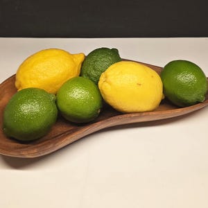 May include: A wooden, leaf-shaped bowl filled with fresh citrus fruits. The arrangement includes bright yellow lemons and vibrant green limes, creating a colorful display. The bowl's natural wood grain adds a rustic touch to the presentation.