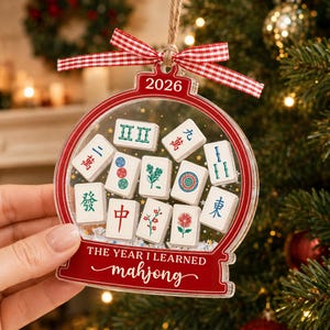 K&ouml;nnte beinhalten: Ein durchsichtiger, kugelf&ouml;rmiger Acryl-Weihnachtsbaumschmuck mit rotem Rand, rot-wei&szlig;em Vichy-Karo-Band und dem Text 2026 sowie THE YEAR I LEARNED mahjong. In der Mitte sind verschiedene wei&szlig;e Mahjong-Steine mit bunten Symbolen abgebildet.