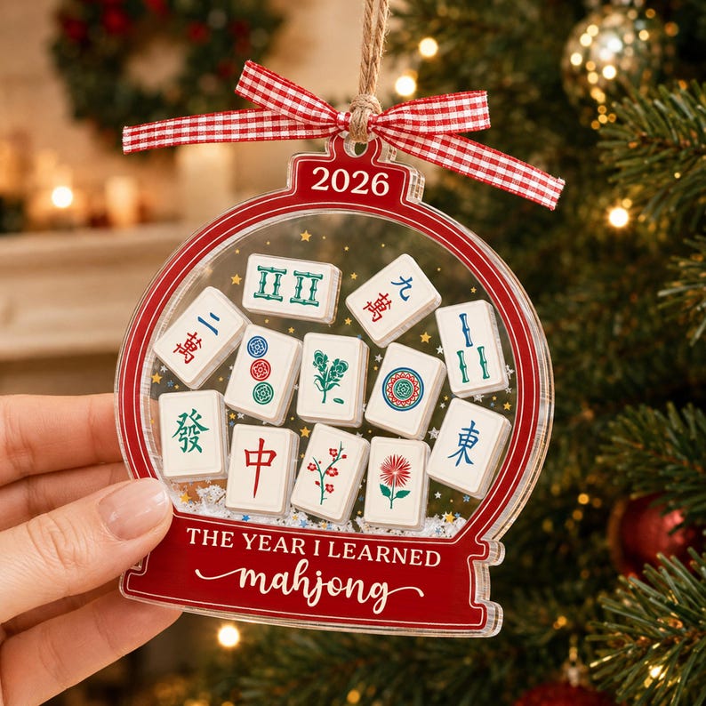 K&ouml;nnte beinhalten: Ein durchsichtiger, kugelf&ouml;rmiger Acryl-Weihnachtsbaumschmuck mit rotem Rand, rot-wei&szlig;em Vichy-Karo-Band und dem Text 2026 sowie THE YEAR I LEARNED mahjong. In der Mitte sind verschiedene wei&szlig;e Mahjong-Steine mit bunten Symbolen abgebildet.