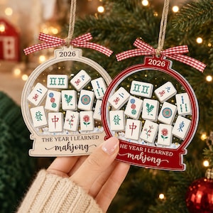 K&ouml;nnte beinhalten: Zwei schneekugelartige Ornamente aus Holz, eines naturfarben und eines rot, mit Mahjong-Spielsteinen im Inneren. Jedes Ornament ist mit einem rot-wei&szlig;en Vichy-Karo-Band versehen und zeigt den Text 2026 sowie THE YEAR I LEARNED mahjong.