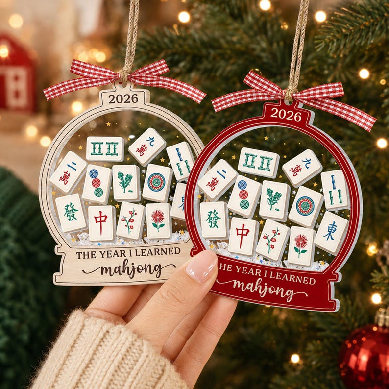 K&ouml;nnte beinhalten: Zwei schneekugelartige Ornamente aus Holz, eines naturfarben und eines rot, mit Mahjong-Spielsteinen im Inneren. Jedes Ornament ist mit einem rot-wei&szlig;en Vichy-Karo-Band versehen und zeigt den Text 2026 sowie THE YEAR I LEARNED mahjong.