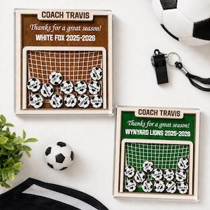May include: Two square wooden display frames featuring a soccer goal graphic with small soccer ball tokens labeled with names and numbers. Each frame is engraved with COACH TRAVIS, Thanks for a great season!, and team names WHITE FOX or WYNYARD LIONS 2025-2026.