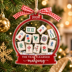 K&ouml;nnte beinhalten: Ein transparenter, kugelf&ouml;rmiger Weihnachtsschmuck mit rotem Rand, einem rot-wei&szlig; karierten Band und dem Text 2026 sowie THE YEAR I LEARNED mahjong. Im Inneren befinden sich verschiedene Mahjong-Steine mit bunten Symbolen.