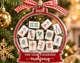 May include: A clear, globe-shaped Christmas ornament featuring a red border, a red and white gingham ribbon, and the text 2026 and THE YEAR I LEARNED mahjong. Inside are various mahjong tiles with colorful symbols and floral designs.