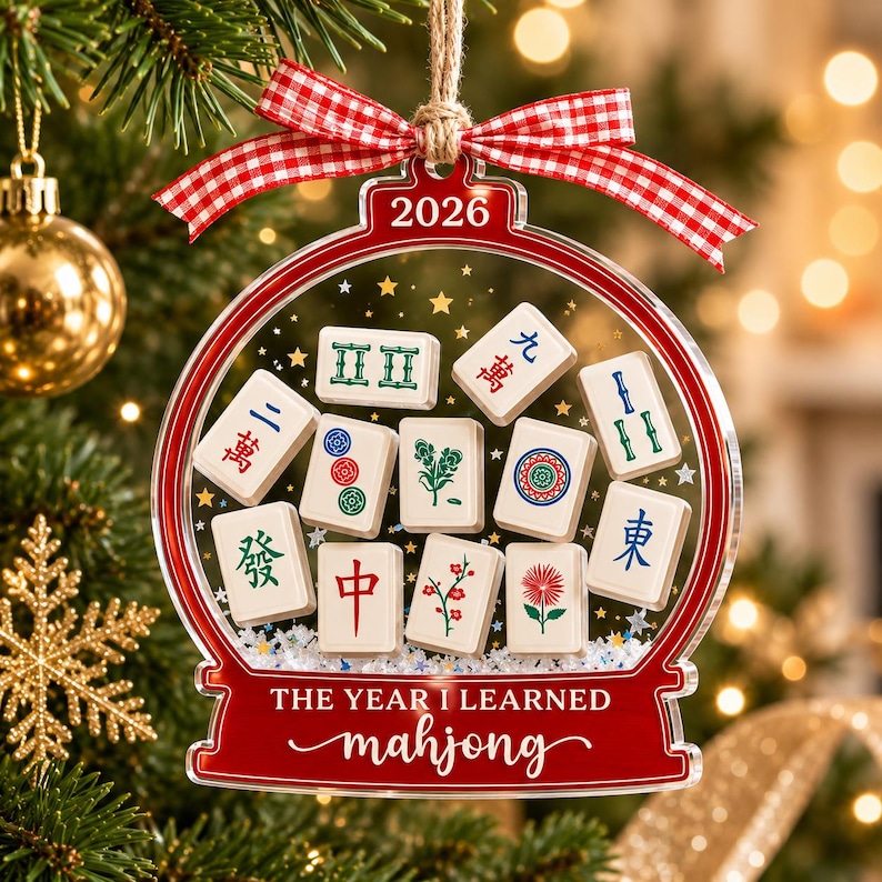 K&ouml;nnte beinhalten: Ein transparenter, kugelf&ouml;rmiger Weihnachtsschmuck mit rotem Rand, einem rot-wei&szlig; karierten Band und dem Text 2026 sowie THE YEAR I LEARNED mahjong. Im Inneren befinden sich verschiedene Mahjong-Steine mit bunten Symbolen.