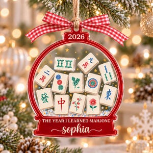 K&ouml;nnte beinhalten: Ein transparenter, kugelf&ouml;rmiger Weihnachtsschmuck mit rotem Rand und einer rot-wei&szlig; karierten Schleife. Er zeigt Mahjong-Steine, goldene Sterne sowie den Text 2026, THE YEAR I LEARNED MAHJONG und den Namen Sophia in wei&szlig;er Schrift.
