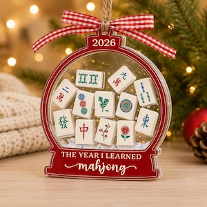 K&ouml;nnte beinhalten: Ein durchsichtiges, kugelf&ouml;rmiges Acryl-Ornament mit rotem Rand, rot-wei&szlig;em Vichy-Karo-Band und dem Text 2026 sowie THE YEAR I LEARNED mahjong. Im Inneren befinden sich wei&szlig;e Mahjong-Steine mit bunten Symbolen und kleinen goldenen Sternen.