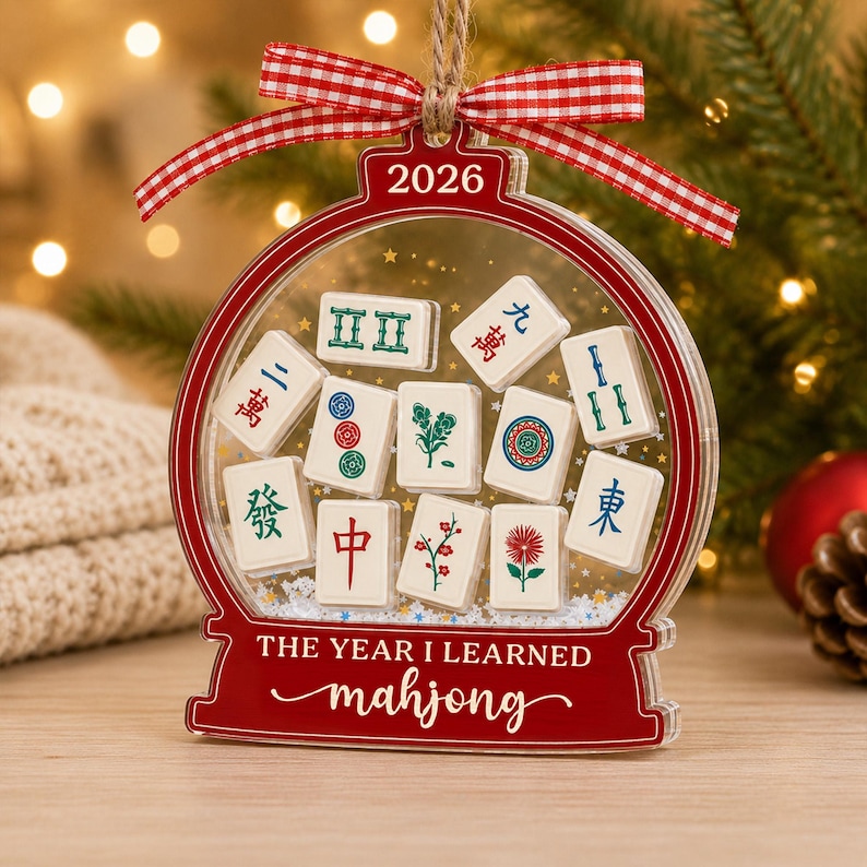 K&ouml;nnte beinhalten: Ein durchsichtiges, kugelf&ouml;rmiges Acryl-Ornament mit rotem Rand, rot-wei&szlig;em Vichy-Karo-Band und dem Text 2026 sowie THE YEAR I LEARNED mahjong. Im Inneren befinden sich wei&szlig;e Mahjong-Steine mit bunten Symbolen und kleinen goldenen Sternen.