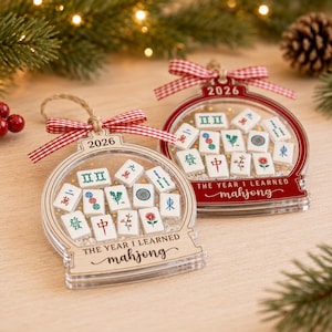 K&ouml;nnte beinhalten: Zwei schneekugelartige Ornamente mit Mahjong-Spielsteinen im Inneren. Eines ist aus Naturholz, das andere ist rot. Beide sind mit 2026 und dem Text THE YEAR I LEARNED mahjong beschriftet und mit einer rot-wei&szlig;en Vichy-Schleife versehen.