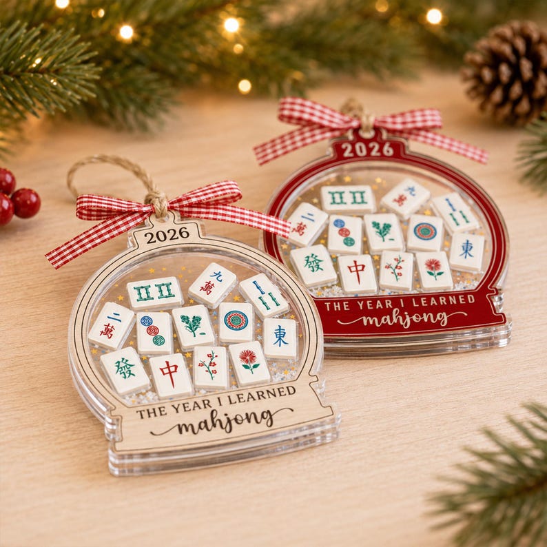 K&ouml;nnte beinhalten: Zwei schneekugelartige Ornamente mit Mahjong-Spielsteinen im Inneren. Eines ist aus Naturholz, das andere ist rot. Beide sind mit 2026 und dem Text THE YEAR I LEARNED mahjong beschriftet und mit einer rot-wei&szlig;en Vichy-Schleife versehen.