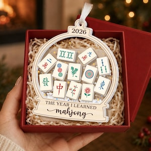 K&ouml;nnte beinhalten: Ein h&ouml;lzernes Schneekugel-Ornament mit dem Text 2026 und THE YEAR I LEARNED mahjong. Das Ornament enth&auml;lt verschiedene wei&szlig;e Mahjong-Steine mit bunten Symbolen und Schriftzeichen und ist an einem wei&szlig;en Band in einer roten Schachtel befestigt.