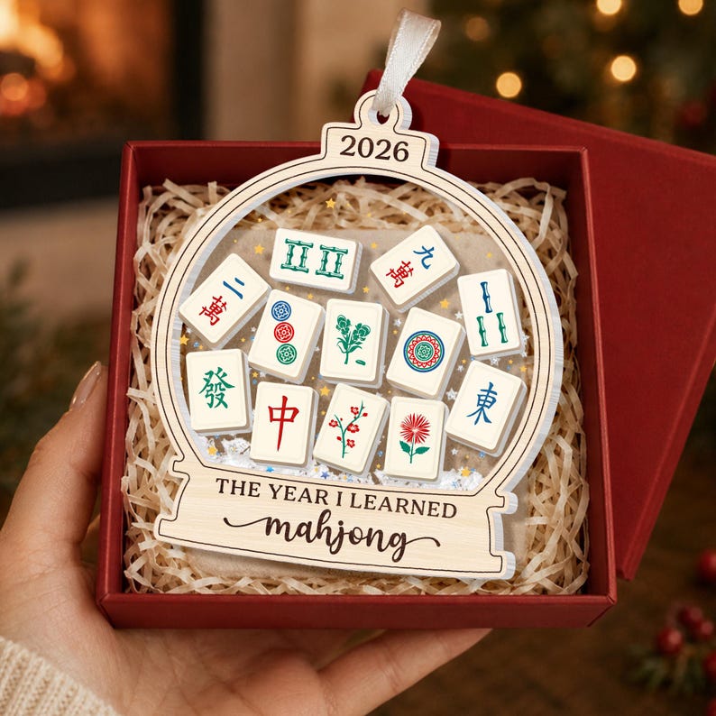 K&ouml;nnte beinhalten: Ein h&ouml;lzernes Schneekugel-Ornament mit dem Text 2026 und THE YEAR I LEARNED mahjong. Das Ornament enth&auml;lt verschiedene wei&szlig;e Mahjong-Steine mit bunten Symbolen und Schriftzeichen und ist an einem wei&szlig;en Band in einer roten Schachtel befestigt.