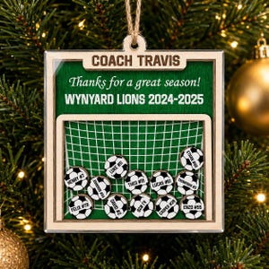 May include: A square wooden ornament featuring a green soccer field background with a white net graphic. Text reads COACH TRAVIS, Thanks for a great season!, and WYNYARD LIONS 2024-2025. Twelve small wooden soccer ball tokens display names and numbers.
