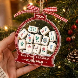 K&ouml;nnte beinhalten: Ein transparenter, kugelf&ouml;rmiger Weihnachtsbaumschmuck mit rotem Rand, rot-wei&szlig;em Vichy-Band und dem Text 2026 sowie THE YEAR I LEARNED mahjong. Im Inneren befinden sich wei&szlig;e Mahjong-Steine mit bunten Symbolen und glitzerndem Kunstschnee.