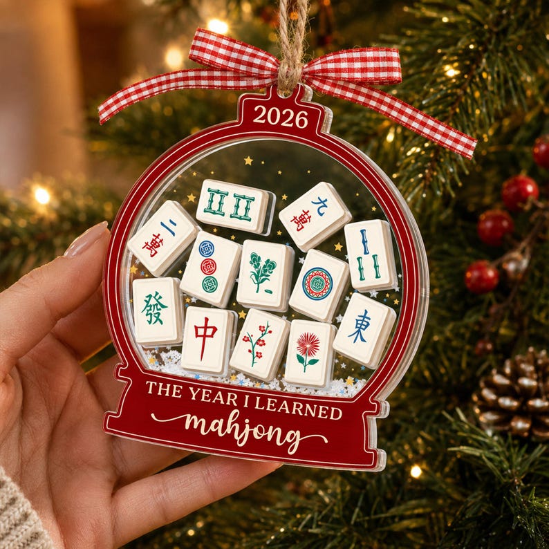 K&ouml;nnte beinhalten: Ein transparenter, kugelf&ouml;rmiger Weihnachtsbaumschmuck mit rotem Rand, rot-wei&szlig;em Vichy-Band und dem Text 2026 sowie THE YEAR I LEARNED mahjong. Im Inneren befinden sich wei&szlig;e Mahjong-Steine mit bunten Symbolen und glitzerndem Kunstschnee.