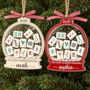 K&ouml;nnte beinhalten: Zwei schneekugelartige Ornamente mit roten Vichy-Schleifen. Eines ist aus hellem Holz, das andere ist rot. Beide zeigen Mahjong-Steine, den Text 2026, THE YEAR I LEARNED MAHJONG sowie die Namen Noah oder Sophia an Schn&uuml;ren.