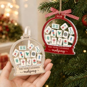 K&ouml;nnte beinhalten: Zwei schneekugelartige Ornamente mit Mahjong-Spielsteinen im Inneren. Eines ist transparent mit einem holzfarbenen Sockel, das andere ist rot. Beide tragen den Text 2026 und THE YEAR I LEARNED mahjong. Das rote Ornament hat ein rot-wei&szlig;es Vichy-Band.