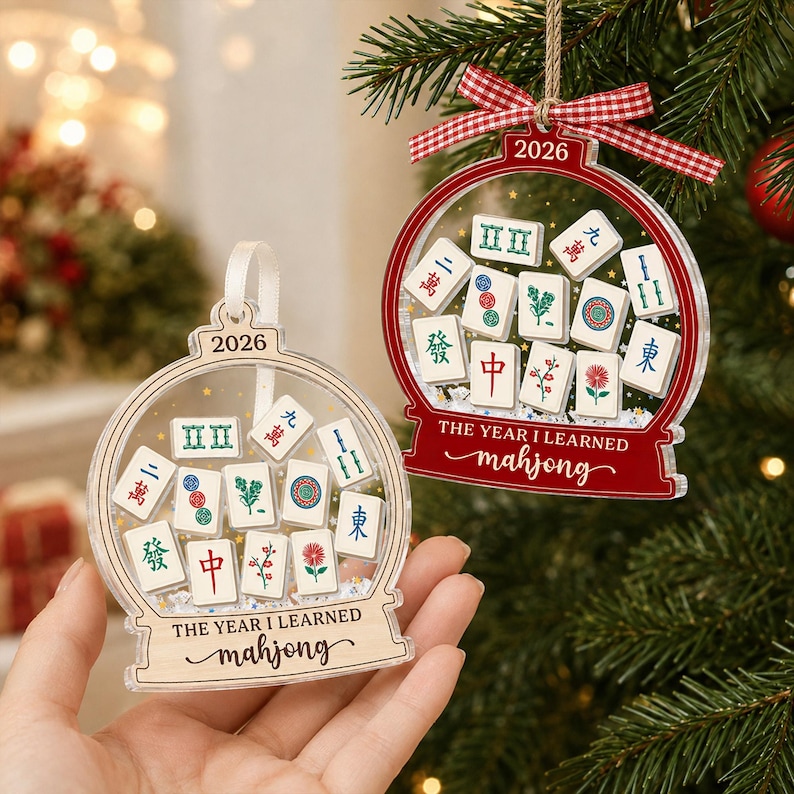 K&ouml;nnte beinhalten: Zwei schneekugelartige Ornamente mit Mahjong-Spielsteinen im Inneren. Eines ist transparent mit einem holzfarbenen Sockel, das andere ist rot. Beide tragen den Text 2026 und THE YEAR I LEARNED mahjong. Das rote Ornament hat ein rot-wei&szlig;es Vichy-Band.