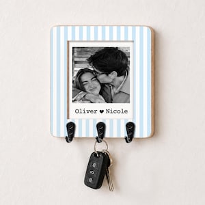 May include: A square wall-mounted key holder featuring a light blue and white vertical striped border. The center displays a black and white photograph of two people with the printed text Oliver heart Nicole. Three black metal hooks hold a car key fob.
