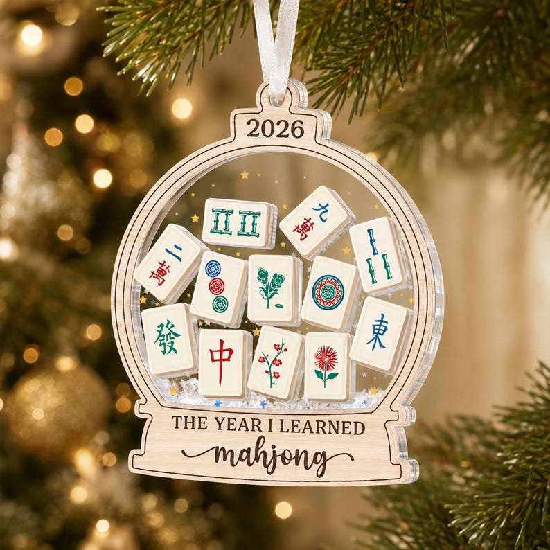 K&ouml;nnte beinhalten: Ein durchsichtiger, schneekugel-f&ouml;rmiger Acryl-Weihnachtsbaumschmuck mit verschiedenen Mahjong-Steinen im Inneren. Oben ist 2026 eingraviert, und der Sockel tr&auml;gt den Text "THE YEAR I LEARNED mahjong" in schwarzer Schrift.