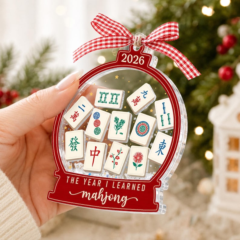 K&ouml;nnte beinhalten: Ein durchsichtiger Acryl-Weihnachtsbaumschmuck in Schneekugelform mit rotem Rand und Mahjong-Spielsteinen im Inneren. Oben steht die Jahreszahl 2026, unten der Text THE YEAR I LEARNED mahjong. Ein rot-wei&szlig; kariertes Band ist oben befestigt.