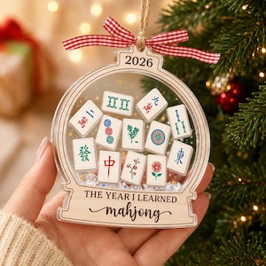 K&ouml;nnte beinhalten: Ein transparenter, kugelf&ouml;rmiger Weihnachtsschmuck mit einer Sammlung von Miniatur-Mahjong-Steinen im Inneren. Oben ist 2026 eingraviert, unten steht THE YEAR I LEARNED mahjong. Der Schmuck ist mit einem rot-wei&szlig;en Vichy-Karo-Band versehen.