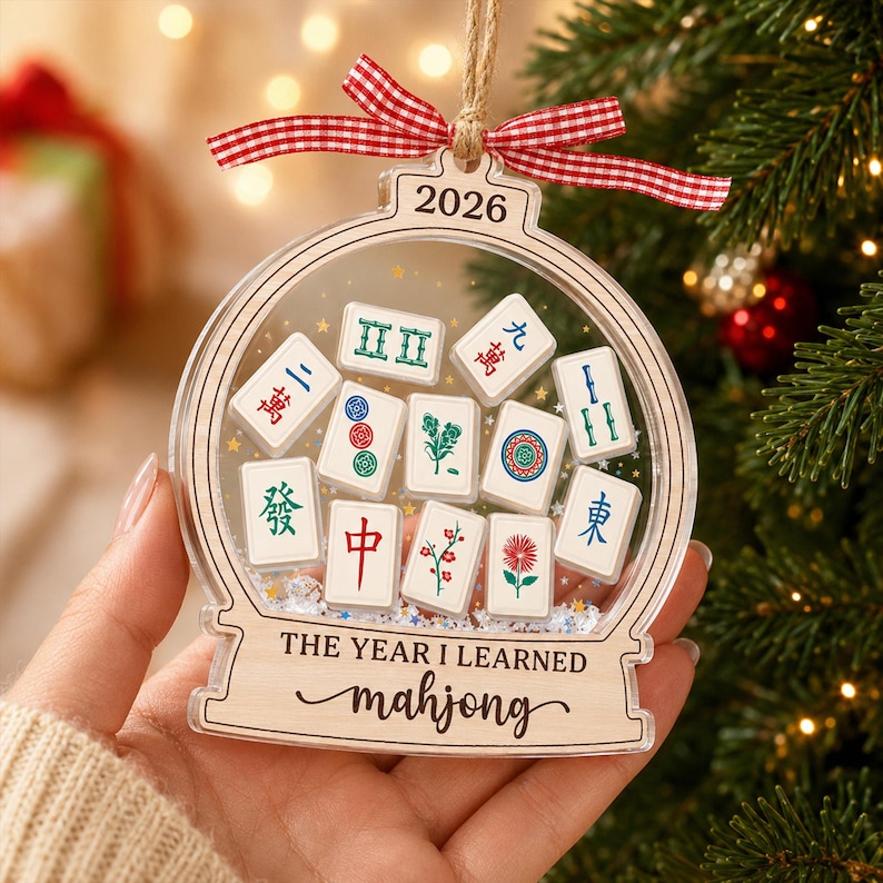 K&ouml;nnte beinhalten: Ein transparenter, kugelf&ouml;rmiger Weihnachtsschmuck mit einer Sammlung von Miniatur-Mahjong-Steinen im Inneren. Oben ist 2026 eingraviert, unten steht THE YEAR I LEARNED mahjong. Der Schmuck ist mit einem rot-wei&szlig;en Vichy-Karo-Band versehen.