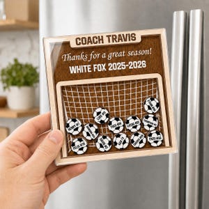 May include: A square wooden plaque featuring a soccer goal graphic with the text COACH TRAVIS, Thanks for a great season!, and WHITE FOX 2025-2026. Eleven small circular soccer ball magnets with names and numbers are attached to the goal net.