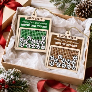 May include: Two square wooden ornaments featuring a soccer goal design with small soccer ball charms inside. One ornament is green with text reading COACH CALEB, Thanks for a great season!, WYNYARD LIONS 2025-2026. The other is brown with COACH TRAVIS, Thanks for a great season!, WHITE FOX 2025-2026.