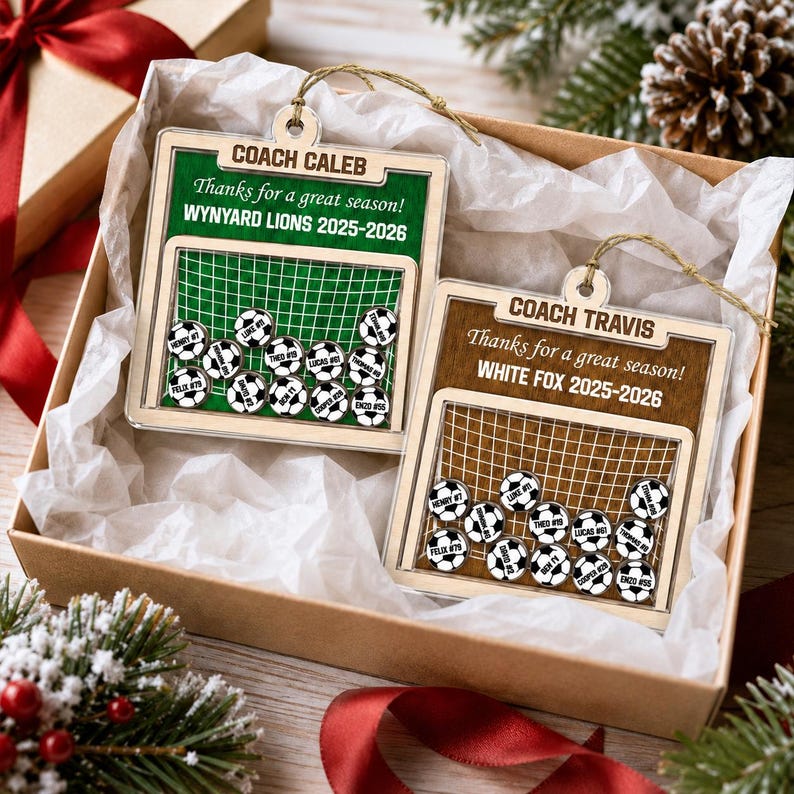 May include: Two square wooden ornaments featuring a soccer goal design with small soccer ball charms inside. One ornament is green with text reading COACH CALEB, Thanks for a great season!, WYNYARD LIONS 2025-2026. The other is brown with COACH TRAVIS, Thanks for a great season!, WHITE FOX 2025-2026.