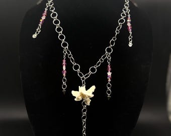 May include: A silver-toned chain necklace featuring two off-white bone vertebrae segments, pink and clear faceted beads, small metal skull charms, and a black heart-shaped pendant. The necklace is displayed on a black stand with a circular logo.