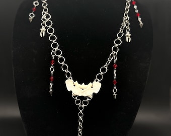 May include: A silver-toned chain necklace featuring a central white bone-like vertebrae segment, dangling red crystal beads, small tooth-shaped charms, and metal spikes. A single tooth charm hangs from a heart-shaped pendant at the bottom center.
