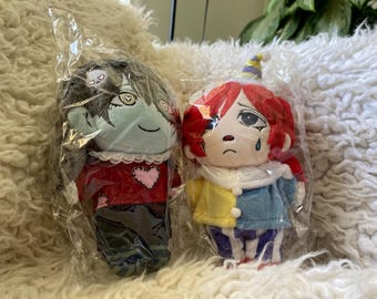 May include: Two plush dolls sealed in clear plastic packaging. The left doll has grey hair, pale blue skin, a red shirt with a pink heart, and a white lace collar. The right doll has red hair, a yellow and blue jacket, and a blue teardrop under one eye.